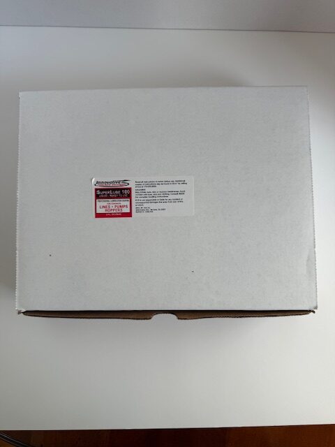White box labeled "Super-Lube 100" with product information sticker, resting on a white surface. The image showcases packaging used for lubricants or similar products.